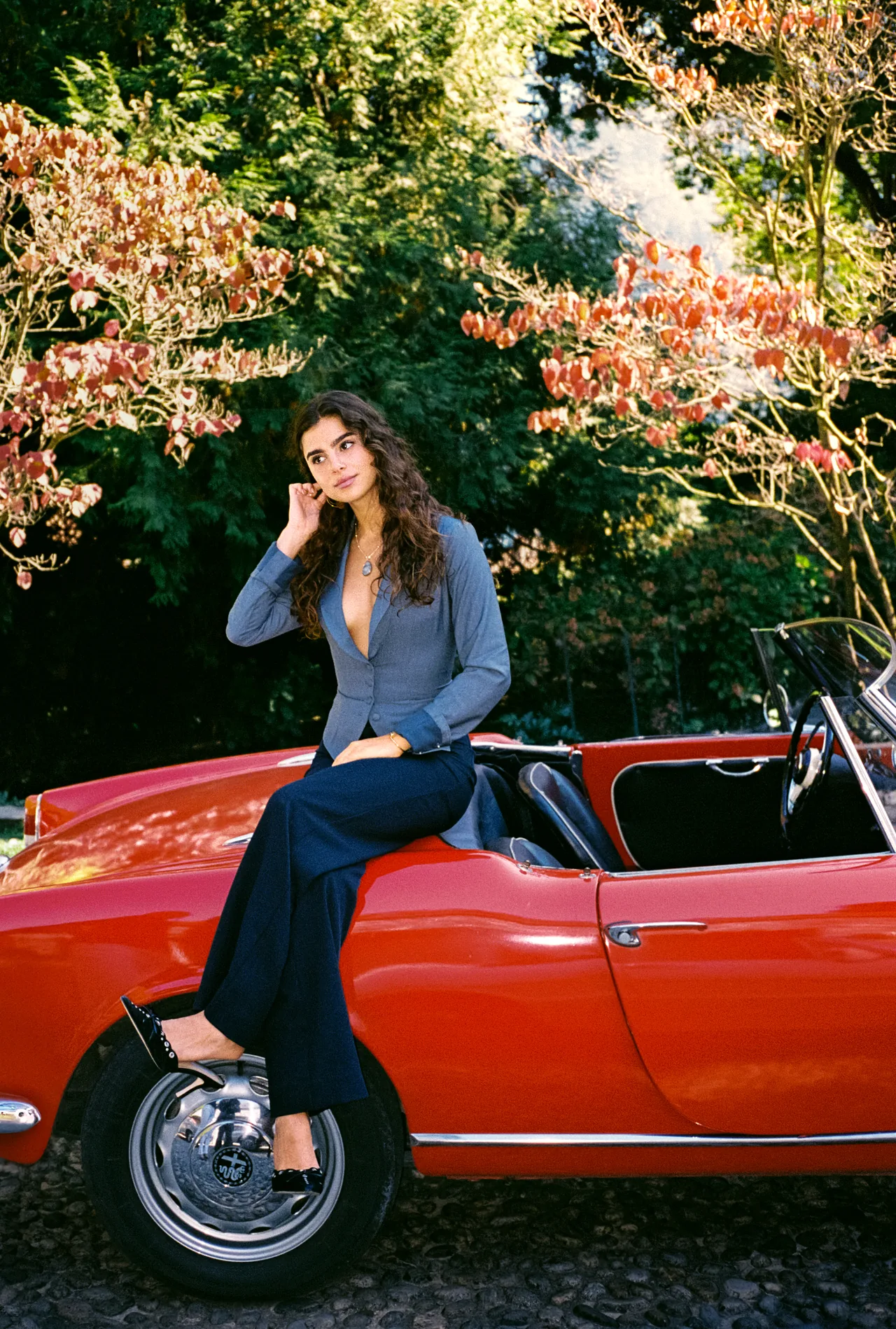 A woman seated on a red classic convertible beneath autumn trees