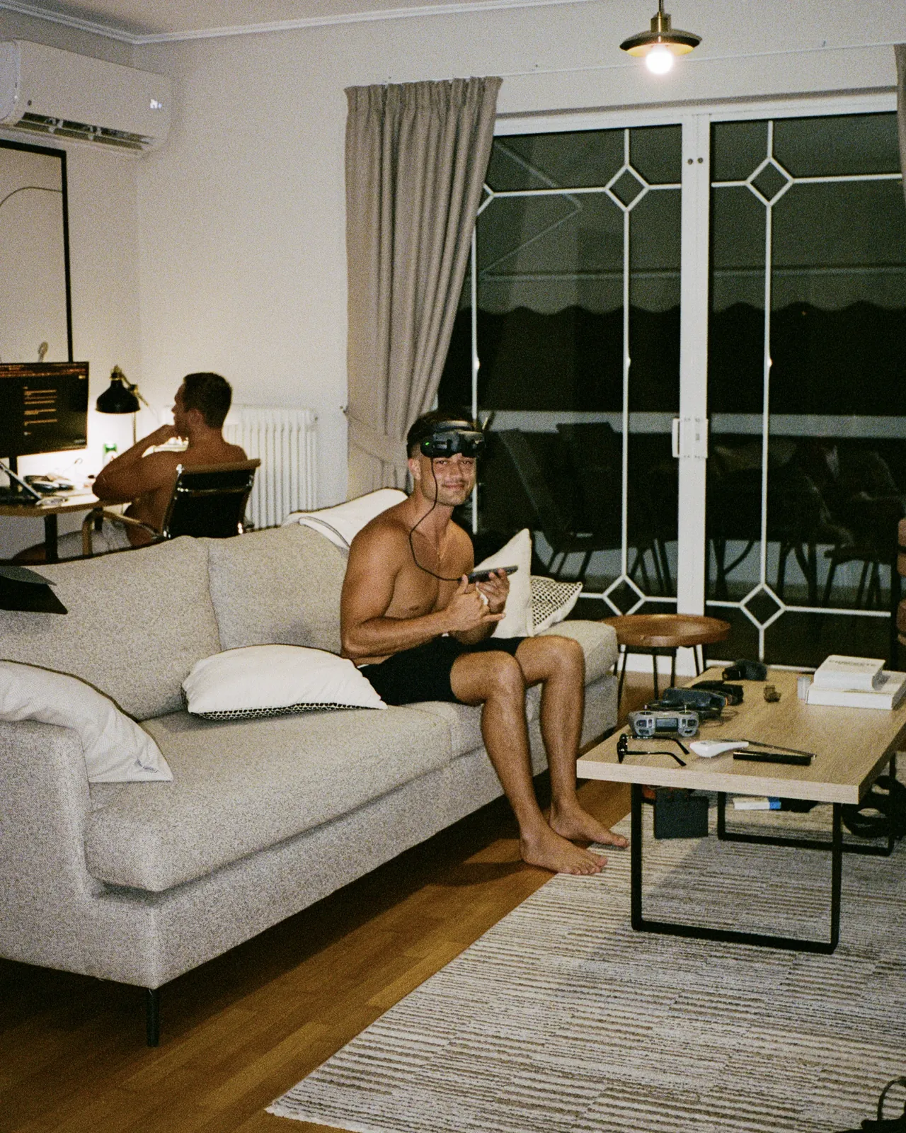 A drone pilot in FPV goggles on a sofa while an editor works behind him