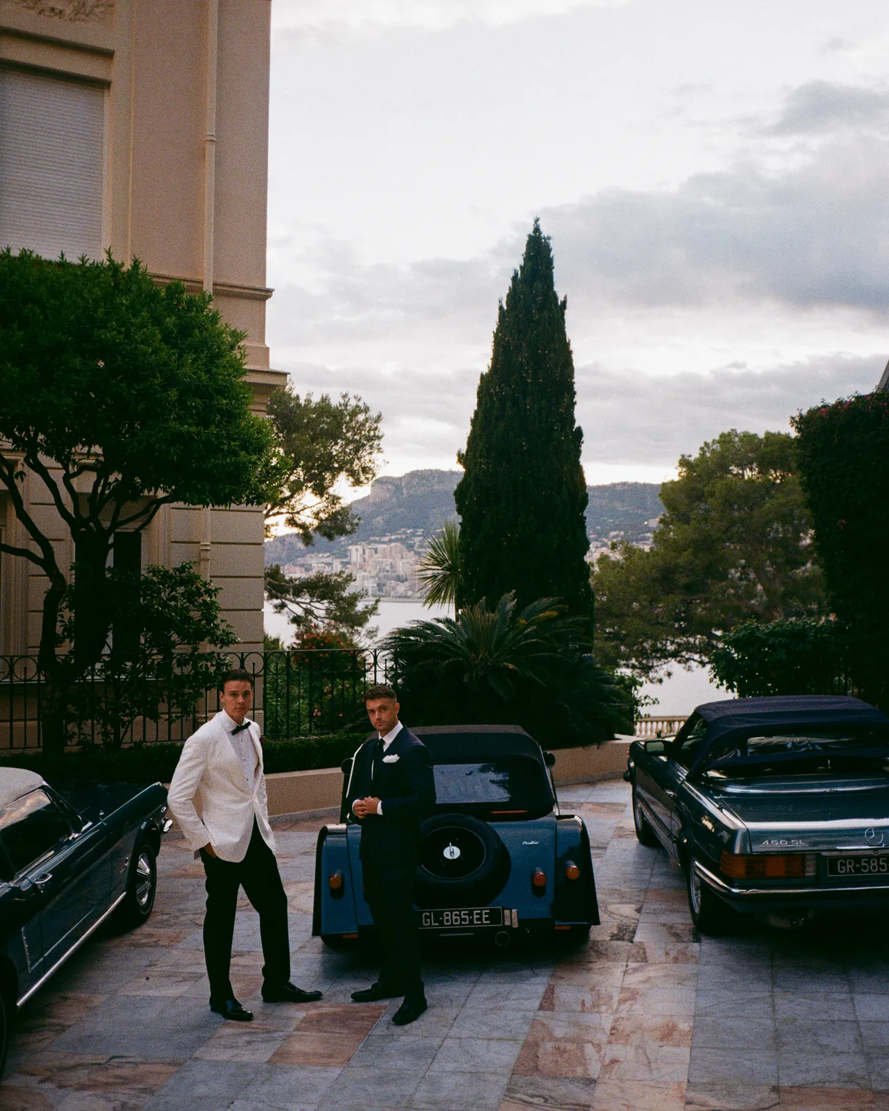 Two men in eveningwear beside classic cars above the coast